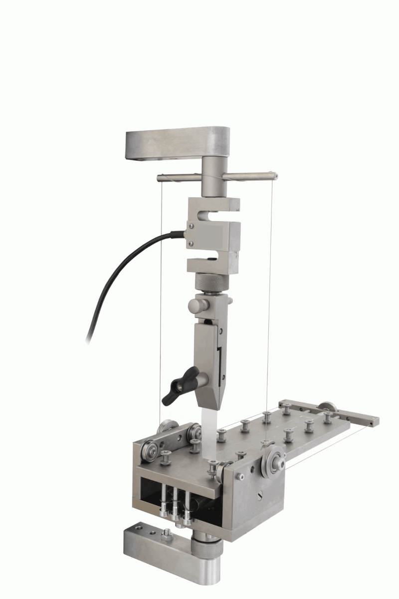 Metallic 90-degree peel test apparatus showing a vertical grip pulling a strip upward while a roller-supported horizontal table moves beneath it to maintain a constant 90-degree peel angle.