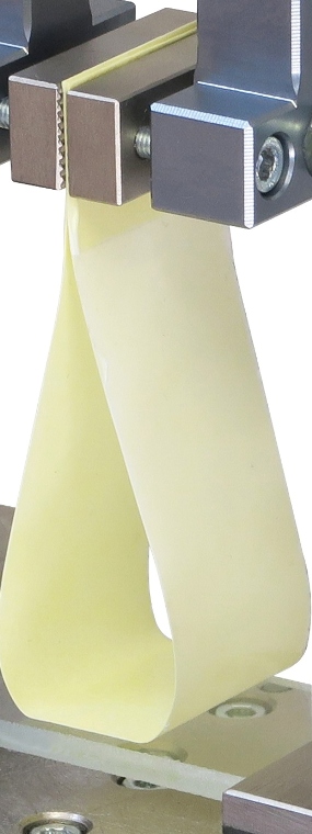 Close-up of a loop tack adhesive test showing a flexible adhesive strip formed into a loop held between two metal grips, used to measure tack strength.