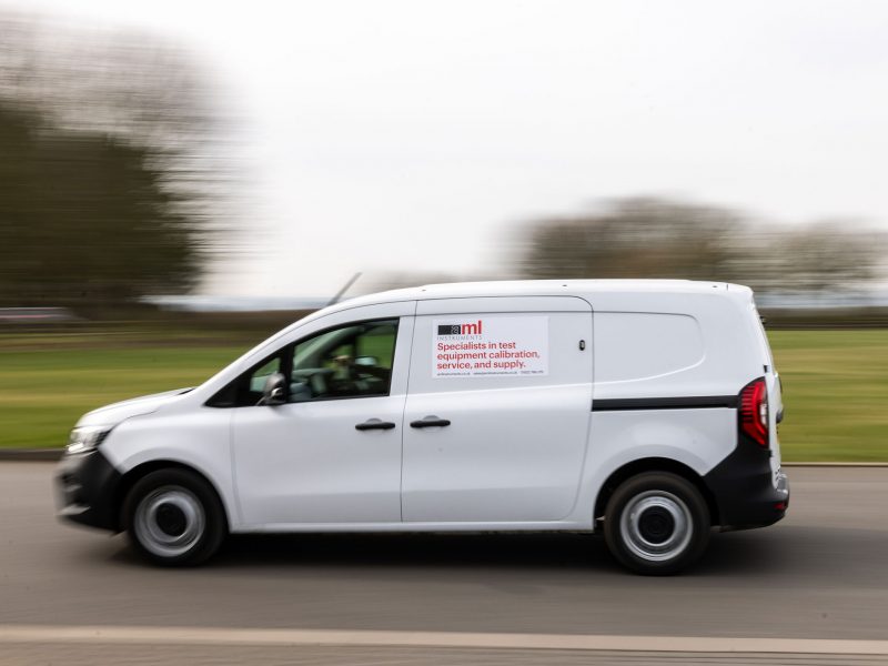 White Ford Transit Connect van with "AML Instruments" branding, parked on grass with trees and cloudy sky in the background.