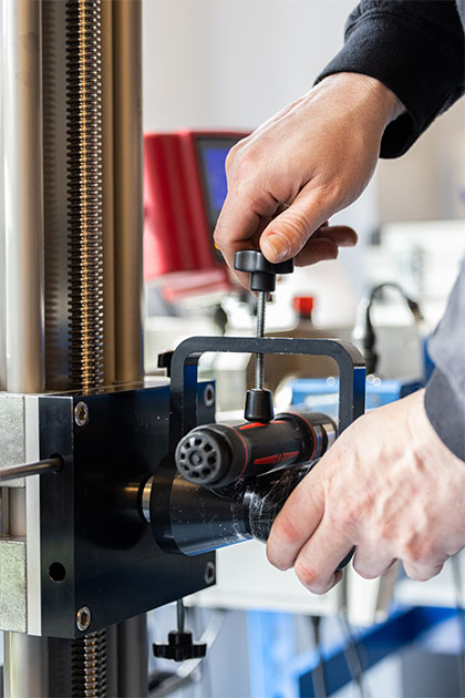 Close-up of hands adjusting a cylindrical component on a piece of torque testing equipment with a vertical metal rail and electronic instruments in the background.